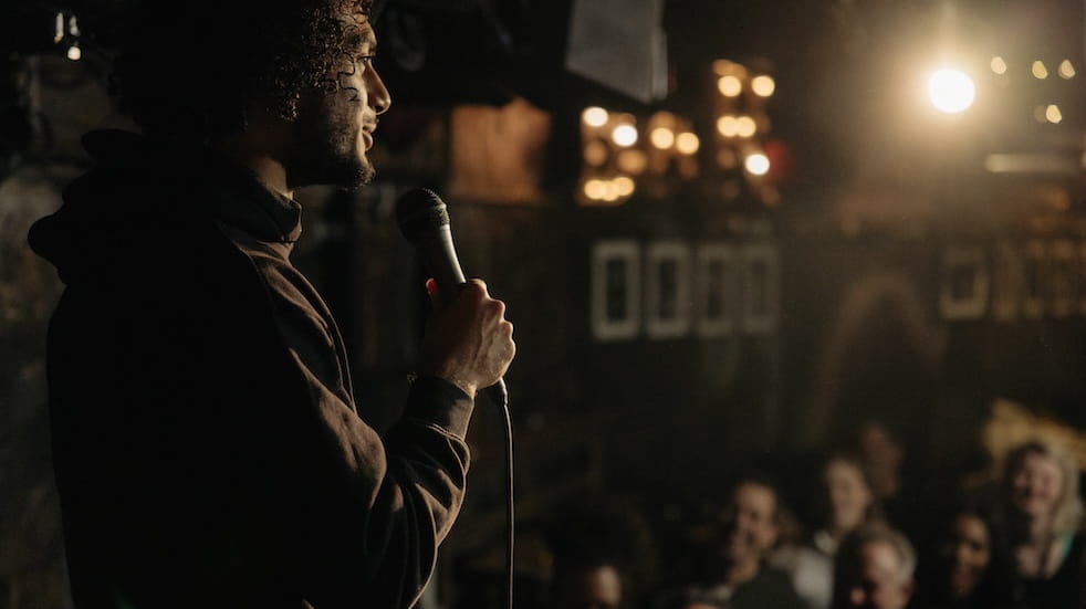 Young male comedian performing stand-up for audience at illuminated amateur theatre